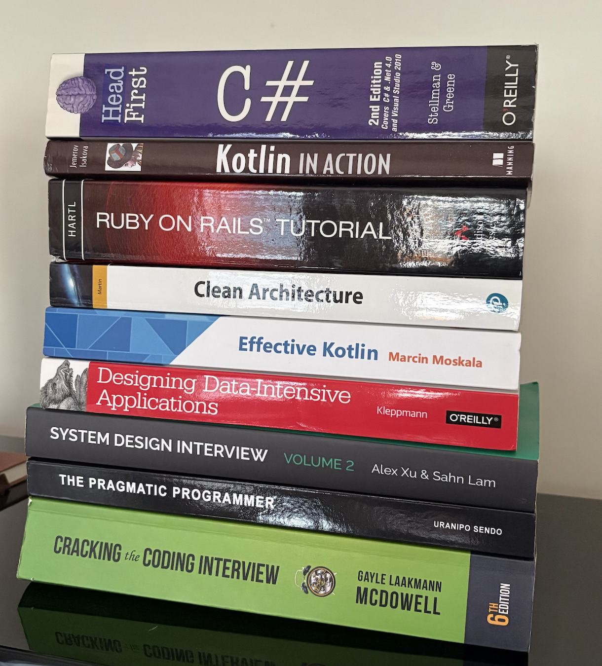 A stack of software engineering books including Head First C#, Kotlin in Action, Ruby on Rails Tutorial, Clean Architecture, Effective Kotlin, Designing Data-Intensive Applications, System Design Interview, The Pragmatic Programmer, and Cracking the Coding Interview.